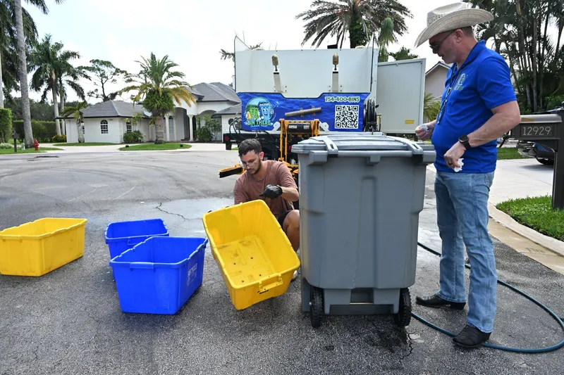 Crew member handling blue and yellow recycling bins beside a gray trash cart