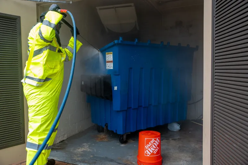 Crew member in high-visibility gear pressure washing a blue dumpster